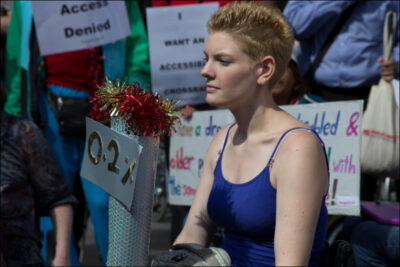 A woman with short blonde hair and a blue vest top looking defiantly forward. A crowd of people are visible behind her and multiple protest signs reading: 