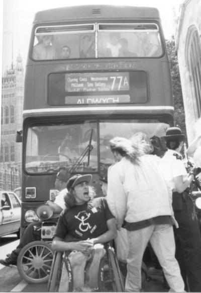 A black and white photo of disabled people blocking a bus. There is a young wheelchair user who is a man sitting in front of the bus. He is shouting something at another man who is facing away fromthe camera. Behind the disabled man you can can make out another wheelchair megaphone on their lap. Behind them is the London bus. It is a 77a going to Aldwych.