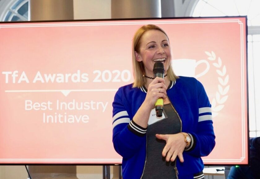 Kirsty, a white woman with blonde hair and wearing a blue jacket, is standing in front of a screen which reads 'TFA Awards 2020' and speaking into a microphone.