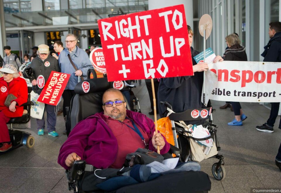 A man using a mobility chair in a train station holds up a protest sign that reads 