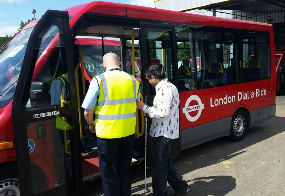 Two people stand by the entrance to a red dial-a-ride bus. On the left is a white man with a bald head, wearing a high-vis jacket. Holding his right elbow is a map with a patterned white shirt and black trousers. This man holds a white cane. Both are facing away from the camera.