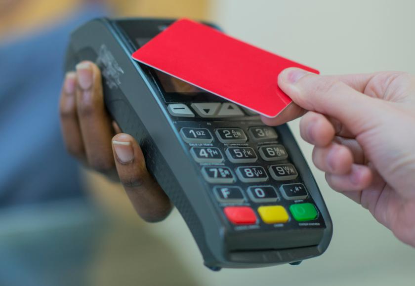 A cardreader is being held out by a person in the distance. Another person is tapping a card onto the reader, as if to pay a bill.