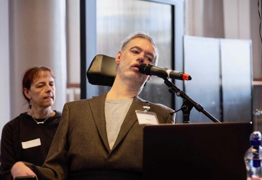 A white man using a mobility chair speaking into a microphone. He is wearing a suit jacket and a name badge.