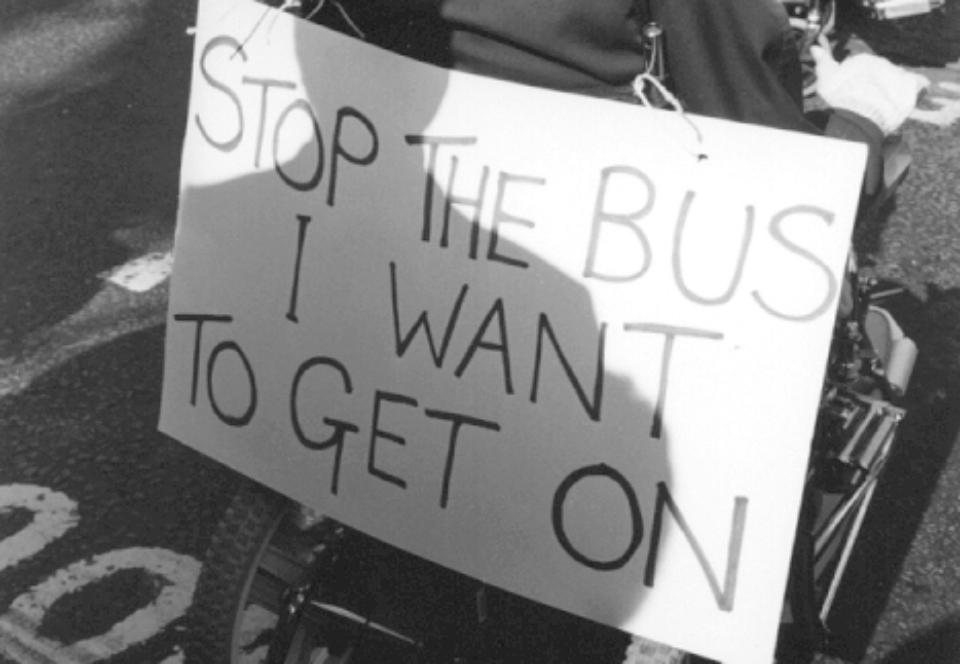 Black and white photo of the back of an electric wheelchair user. They are wearing a black cap. On the back of the bus there is a slogan saying 