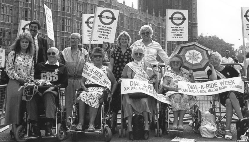 A group of 10 people, some of whom are wheelchair users. They are all standing in a row outside the Houses of Parliament. Some are holding a sign saying 