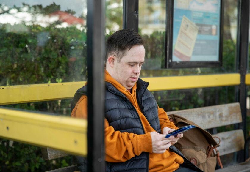 A white man sitting on a bench in a bus stop using a mobile phone. He has short dark brown hair and is wearing an orange hoodie and black gilet.