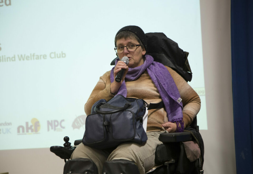 A woman using a mobility chair speaks into a microphone in front of a presentation screen. She is wearing a hat, glasses and a purple scarf.
