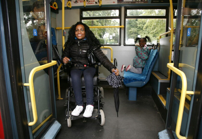 A female wheelchair user on a bus holding a closed umbrella smiles at the camera through the open doors of the bus. She has long black hair, a black jacket and white shoes.