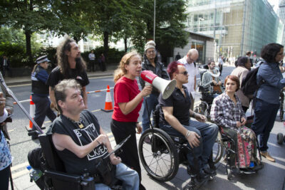 A group of people gathered together on a street with several using wheelchairs. A white woman wearing a red t-shirt stands in the centre speaking into a megaphone.
