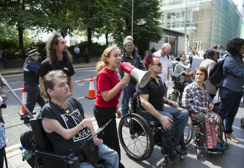 A group of people gathered together on a street with several using wheelchairs. A white woman wearing a red t-shirt stands in the centre speaking into a megaphone.