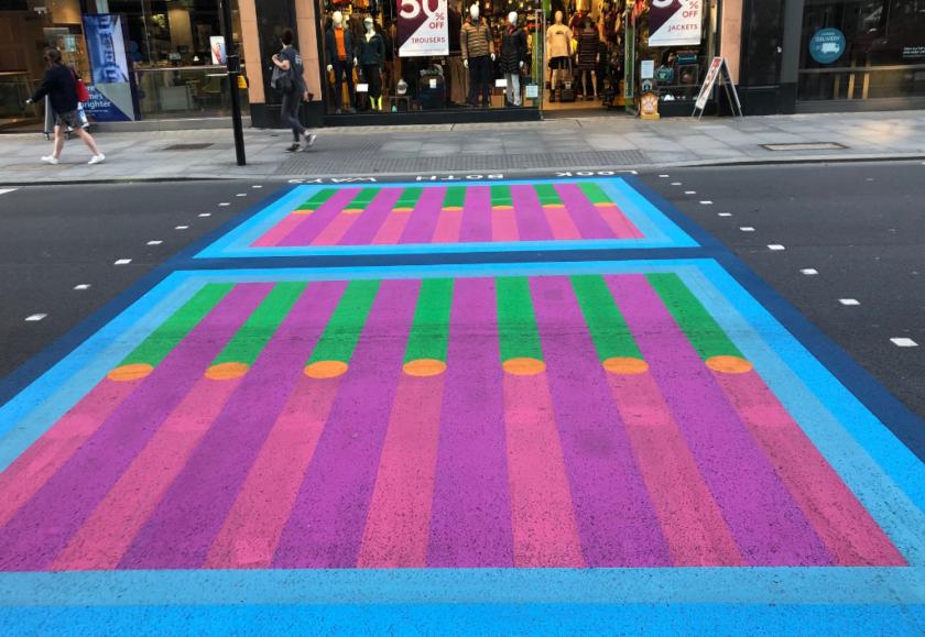 A pedestrian crossing on a high street road is painted with stripes and dots in blue, purple, pink, green and orange.