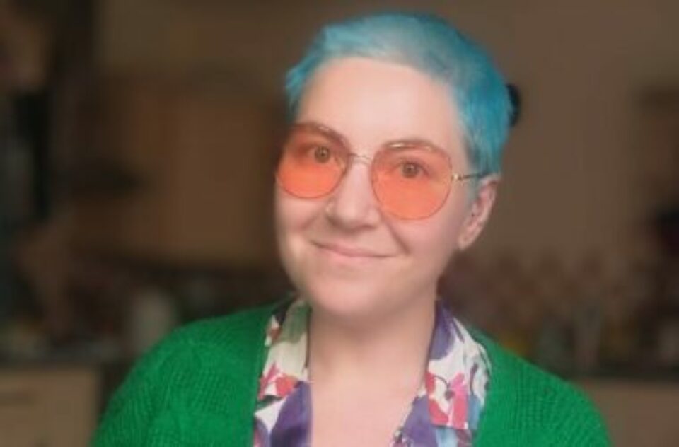 A person with short electric blue hair smiles at the camera. They are wearing pink tinted glasses and a green cardigan.