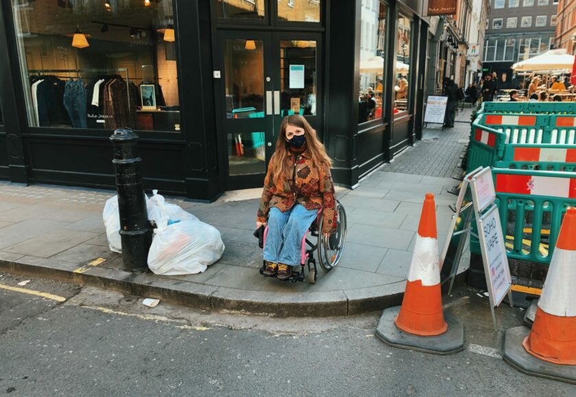 A white woman with long brown hair wearing a protective face mask and using a wheelchair looking into the camera. She is facing the edge of a pavement with no dropped kerb.