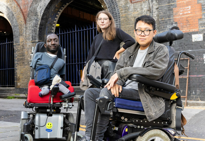 Photograph of three disabled people looking defiantly into the camera. On the left is a black man with upper and lower limb differences who uses an electric wheelchair. He has short brown hair and a beard, and is wearing a blue t-shirt and bag. In the middle is a white woman standing with her arm on her hip. She has brown hair, wears glasses, and a black jumper. On the right is an Asian man who uses an electric wheelchair. He has dark hear, and wears glasses, a grey denim jacket, and jeans.