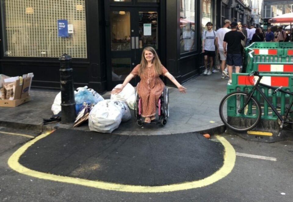 A white woman with long brown hair and a brown dress using a wheelchair smiling at the camera and posing with her arms extended. She is facing the edge of the pavement with a new dropped kerb. Around her are multiple obstacles on a street pavement including several bin bags and boxes.