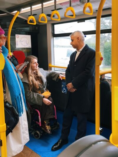 Two people are talking together on a bus, other people are standing nearby. On the left is a woman seated in a bright pink wheelchair. She wears a grey coat and has long brown hair. On the right is Sadiq Khan, Mayor of London. He is standing with his back to the window.