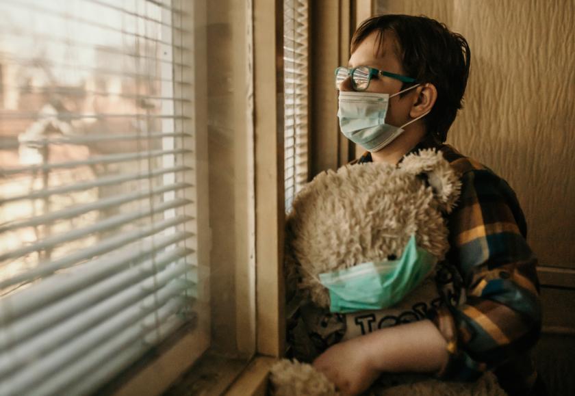 A person with short hair and glasses looking out a window. They are holding a teddy bear and both the person and the teddy are wearing protective face masks.