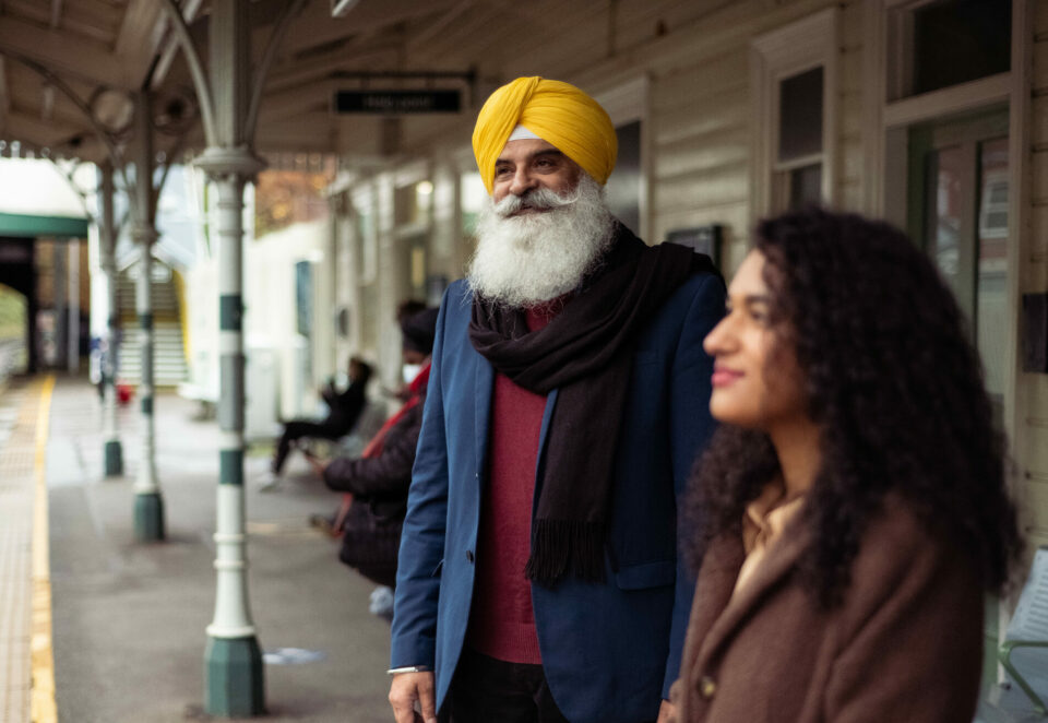 A smiling man and woman stand on a train platform. The man is wearing a yellow turban, blue jacket and scarf and has a white beard and moustache. The woman has brown curls and is wearing a brown jacket.