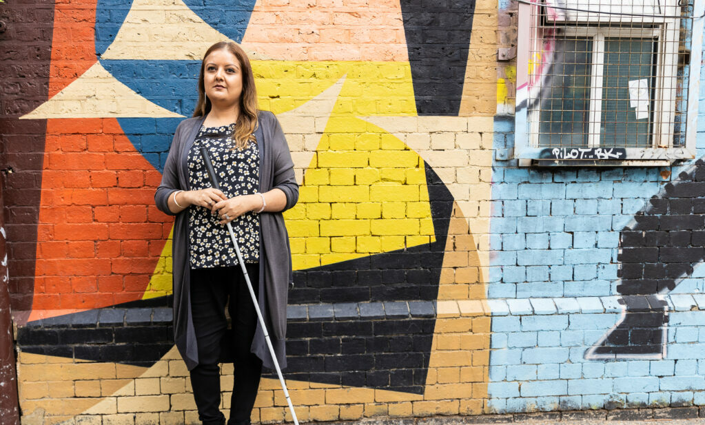 A woman holding a white cane posing in front of a colourful brick wall. She has long brown hair and is wearing a black floral top, long grey cardigan and black trousers.