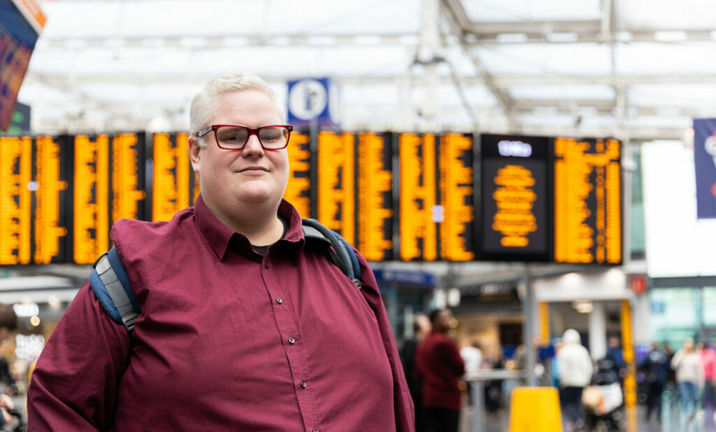 A white man with glasses, short white hair and a red shirt smiling at the camera. He is standing in a train station with information screens in the background.