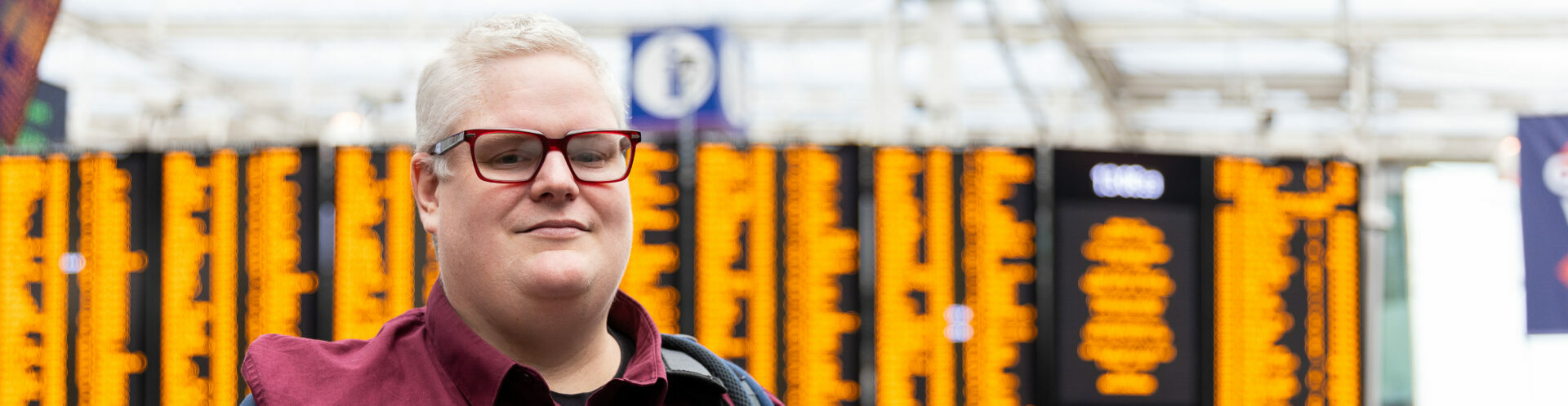 A white man with glasses, short white hair and a red shirt smiling at the camera. He is standing in a train station with information screens in the background.
