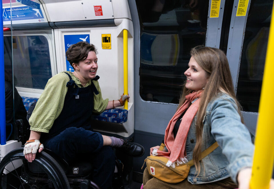 Two people sit together in the wheelchair space of a tube train. On the left is a white person with short brown hair, a green shirt and black dungarees. On the left is a white woman with long brown hair and wearing a denim jacket.