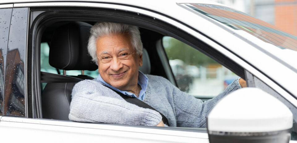An older man with white hair sits behind the wheel of a car. He is smiling at the camera and his elbow rests on the open car window.
