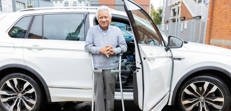An older man with white hair stands by a car with an open driver's door. He is using crutches. He wears a grey jumper and smart trousers.