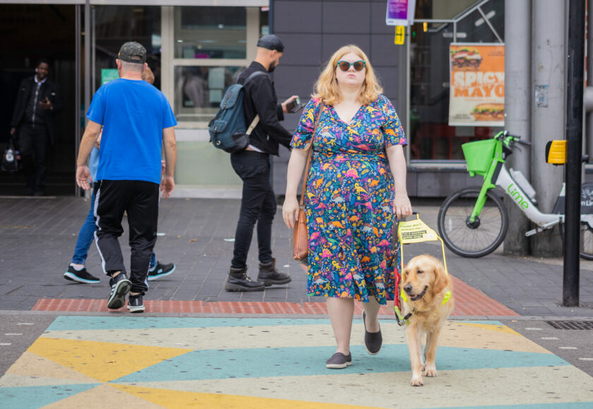 A white woman with blonde hair walks across a road. She wears turquoise sunglasses and a colourful dress with dinosaurs on. She holds the harness of a guide dog who walks beside her.