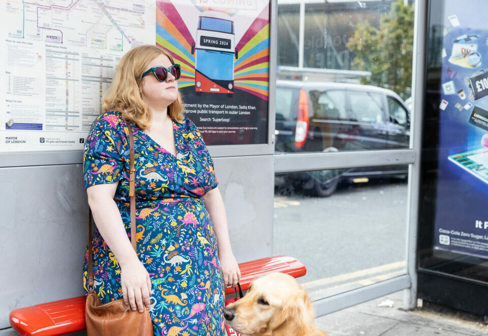 A white woman with blonde hair sits at a bus stop. She wears turquoise sunglasses and a colourful dress with dinosaurs on. She holds the harness of a guide dog who sits beside her.