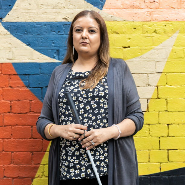 A woman holding a cane is standing in front of a wall which is painted bright colours. She wears a grey cardigan over a patterned blue top.
