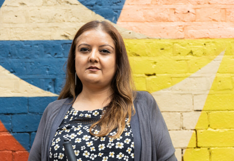 A woman is standing in front of a wall which is painted bright colours. She wears a grey cardigan over a patterned blue top.