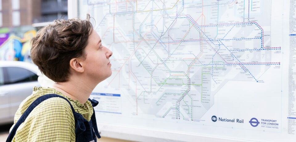 A person looks up at a tube map which is displayed on a board outside a station. They have short brown hair, and wear a green shirt and black dungarees.
