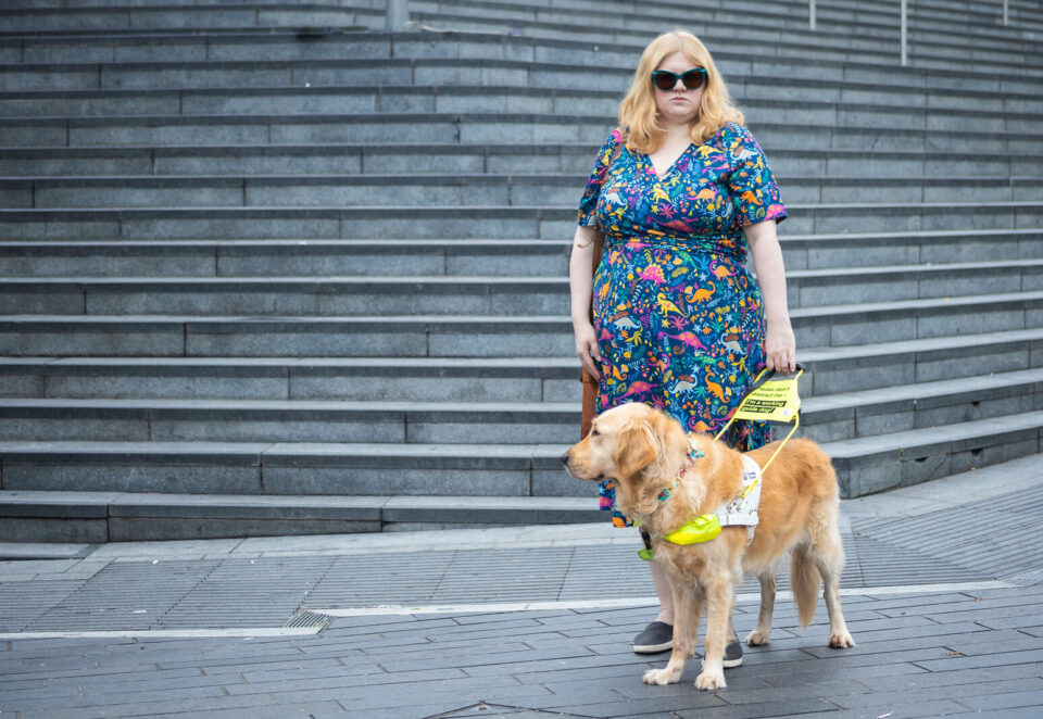 A white woman with blonde hair looks at the camera with a defiant stare. She wears turquoise sunglasses and a colourful dress with dinosaurs on. She holds the harness of a guide dog who stands in front of her.