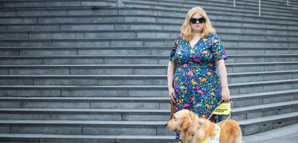 A white woman with blonde hair looks at the camera with a defiant stare. She wears turquoise sunglasses and a colourful dress with dinosaurs on. She holds the harness of a guide dog who stands in front of her.