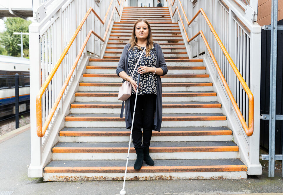 A woman holding a cane is standing on the bottom step of a staircase on a train platform. She looks defiant as she faces the camera. She wears a grey cardigan over a patterned blue top.