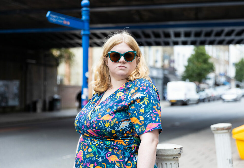 A white woman with blonde hair looks at the camera with a defiant stare. She wears turquoise sunglasses and a colourful dress with dinosaurs on.