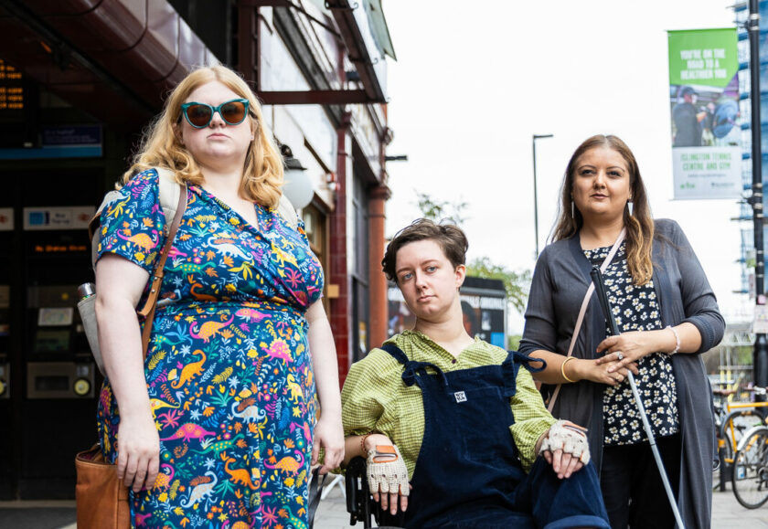 Three disabled people side-by-side in front of a London Underground station. On the left is a woman holding the harness of a guide dog next to her, in the middle is a wheelchair user and on the left is a women using a white cane.