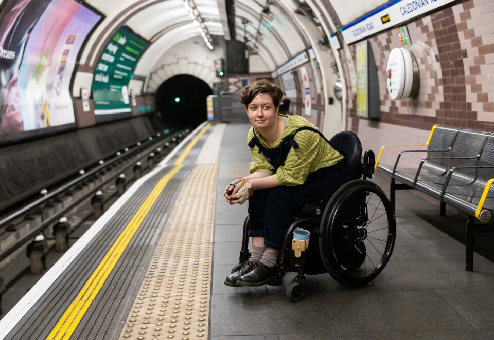 A person using a wheelchair on a tube platform. They are facing the camera but looking away. They have short brown hair, black dungarees and fingerless gloves.