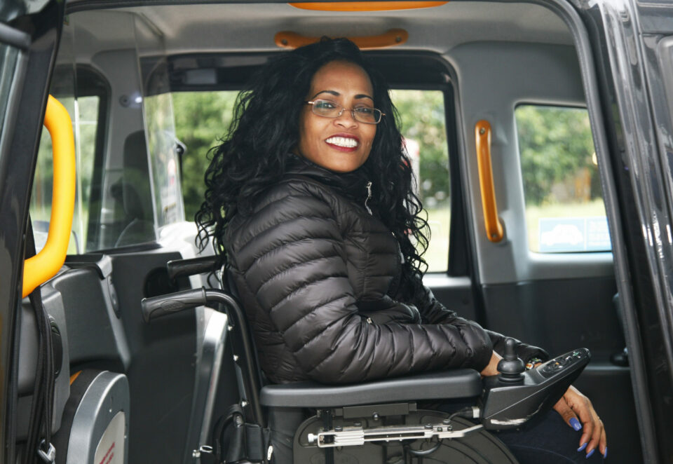A woman sits inside a black cab. She is seated in a wheelchair, and wears a black puffa jacket. She has curly long black hair and is smiling.