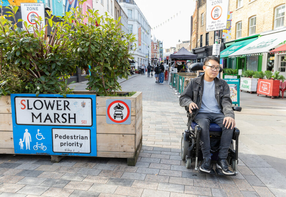 An Asian man who uses an electric wheelchair is in a pedestrianised street. He has dark hair, and wears glasses, a grey denim jacket, and jeans. Behind him are market stalls. On the left is a planter with a sign which reads 'Slower Marsh, pedestrian priority'.