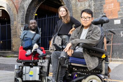 Three disabled people looking defiantly into the camera. On the left is a Black man with upper and lower limb differences who uses an electric wheelchair. He has short brown hair and a beard, and is wearing a blue t-shirt and bag. In the middle is a white woman standing with her arm on her hip. She has brown hair, wears glasses, and a black jumper. On the right is an Asian man who uses an electric wheelchair. He has dark hair, and wears glasses, a grey denim jacket, and jeans.