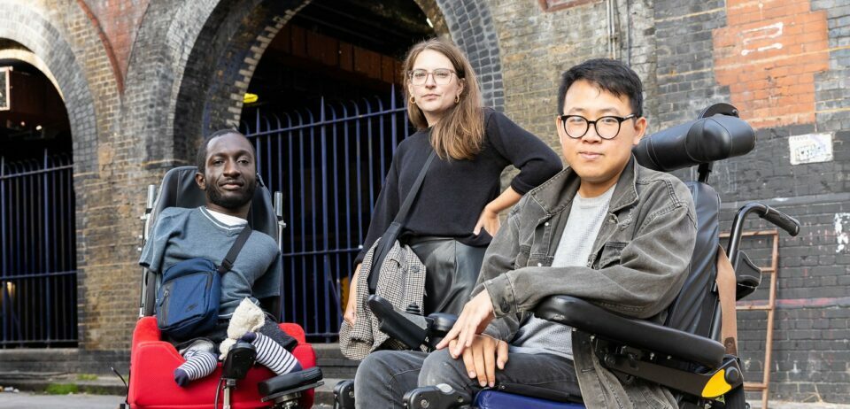 Three disabled people looking defiantly into the camera. On the left is a Black man with upper and lower limb differences who uses an electric wheelchair. He has short brown hair and a beard, and is wearing a blue t-shirt and bag. In the middle is a white woman standing with her arm on her hip. She has brown hair, wears glasses, and a black jumper. On the right is an Asian man who uses an electric wheelchair. He has dark hair, and wears glasses, a grey denim jacket, and jeans.