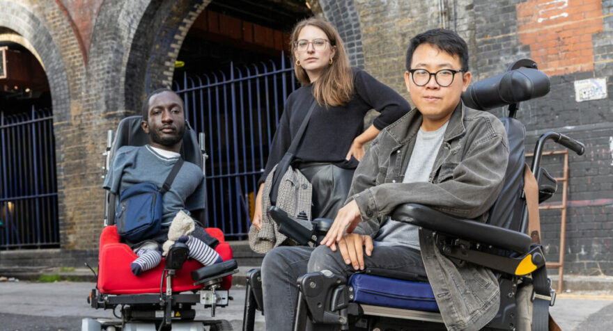 Three disabled people looking defiantly into the camera. On the left is a black man with upper and lower limb differences who uses an electric wheelchair. He has short brown hair and a beard, and is wearing a blue t-shirt and bag. In the middle is a white woman standing with her arm on her hip. She has brown hair, wears glasses, and a black jumper. On the right is an Asian man who uses an electric wheelchair. He has dark hair, and wears glasses, a grey denim jacket, and jeans.