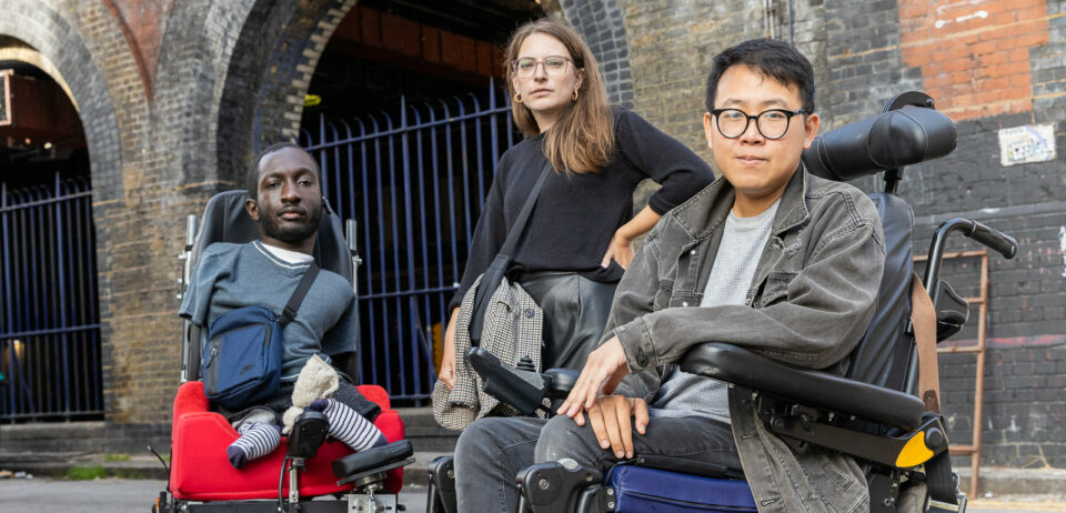 Three disabled people looking defiantly into the camera. On the left is a black man with upper and lower limb differences who uses an electric wheelchair. He has short brown hair and a beard, and is wearing a blue t-shirt and bag. In the middle is a white woman standing with her arm on her hip. She has brown hair, wears glasses, and a black jumper. On the right is an Asian man who uses an electric wheelchair. He has dark hair, and wears glasses, a grey denim jacket, and jeans.