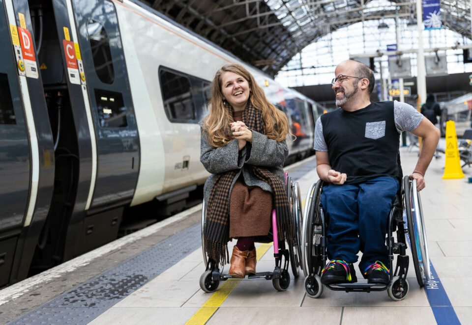 Two people, both wheelchair users, are on the platform at a large train station. On the left is a white woman with long brown hair, wearing a grey coat over a brown dress. On the right is a white man with a beard and glasses, wearing a black t-shirt and blue jeans. They are laughing together.