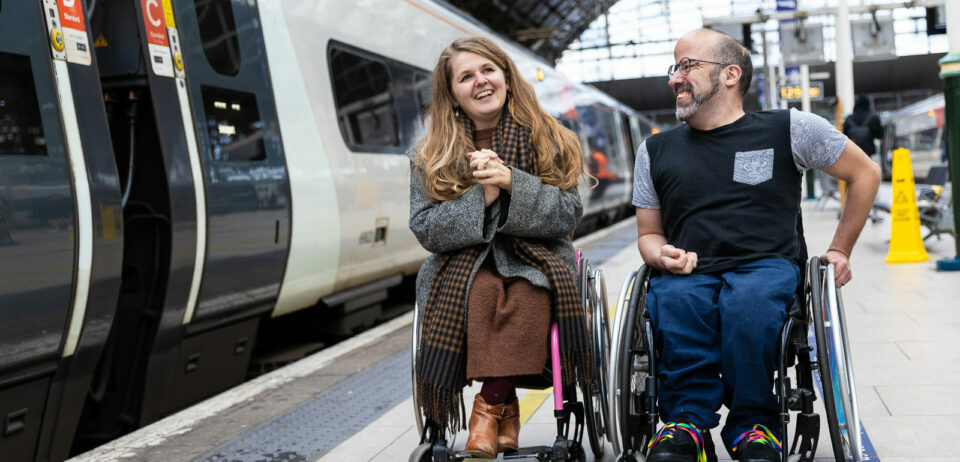 Two people, both wheelchair users, are on the platform at a large train station. On the left is a white woman with long brown hair, wearing a grey coat over a brown dress. On the right is a white man with a beard and glasses, wearing a black t-shirt and blue jeans. They are laughing together.