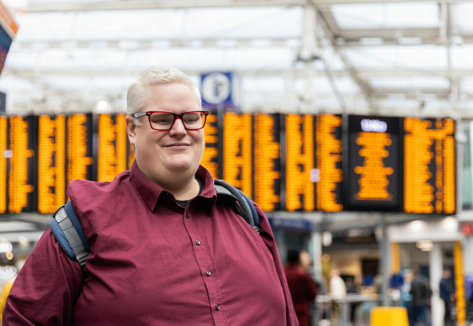 A smiling white man in a train station with train information screens in the background. The man has short white hair, glasses and a red shirt.