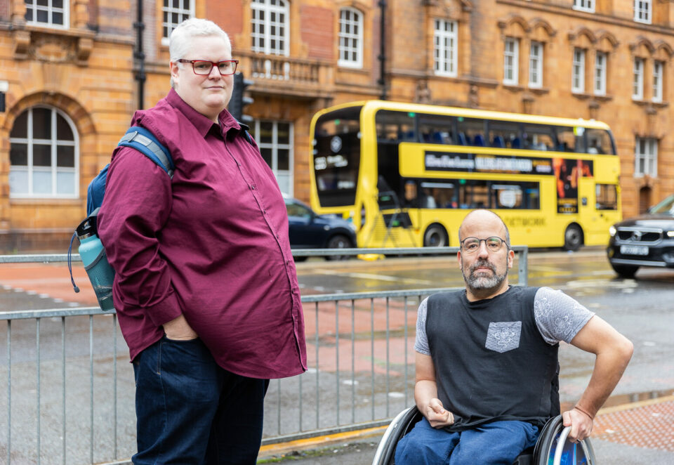 Two white men stand in front of a bright yellow bus in Manchester. On the left is a man with white hair and a deep red shirt. On the right a man with a beard, wearing a black t-shirt.