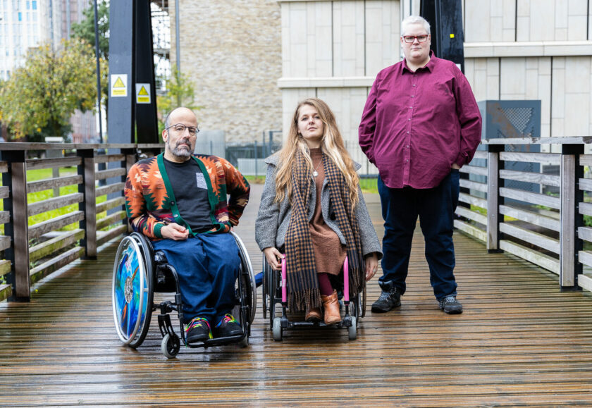 A group of three disabled people face the camera. They are standing on a bridge. On the left is a white man wearing an orange, green and brown cardigan, he is a wheelchair user whose chair has bright blue wheels. In the middle is a white woman with long brown hair, a grey coat and brown dress. Her wheelchair is pink and grey. On the right is a white man standing wearing a deep red shirt.